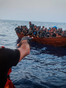 An SMH Rescue crew member in a red helmet and jacket gestures towards an overcrowded wooden boat filled with migrants in the open sea during a rescue operation.
