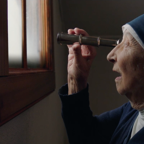 An elderly nun looks through a handheld telescope toward a window, symbolising hope and devotion.