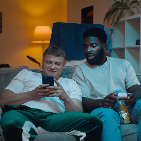 Two men sitting on a sofa at home, one looking at a mobile phone while the other holds a games controller.