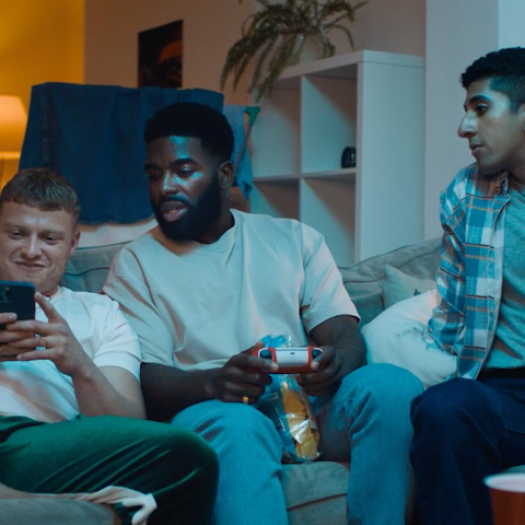 Three men sitting together on a sofa in a living room, watching something on a phone while one holds a games controller.