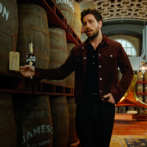 Aaron Taylor-Johnson inside a Jameson distillery surrounded by stacked whiskey barrels, holding a bottle of Jameson Black Barrel.