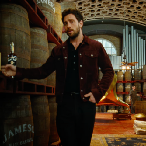 Aaron Taylor-Johnson walking through a Jameson distillery warehouse, touching a whiskey barrel beside a bottle of Jameson Black Barrel.