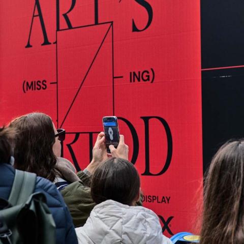 A group of people photographing a large bold red and black outdoor advertisement reading 'Art's (Miss-ing) Period' for Kotex, with one person holding up a smartphone to capture the display. The crowd is seen from behind, wearing winter coats and backpacks.