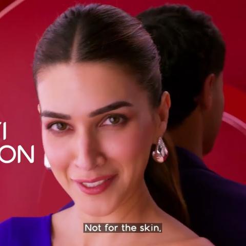 "A still from a Colgate advertisement featuring actor Kriti Sanon, smiling against a bold red background with circular graphic elements. She wears a blue outfit and silver earrings, with her hair pulled back. A second figure is partially visible behind her. On-screen text reads 'Not for the skin'."