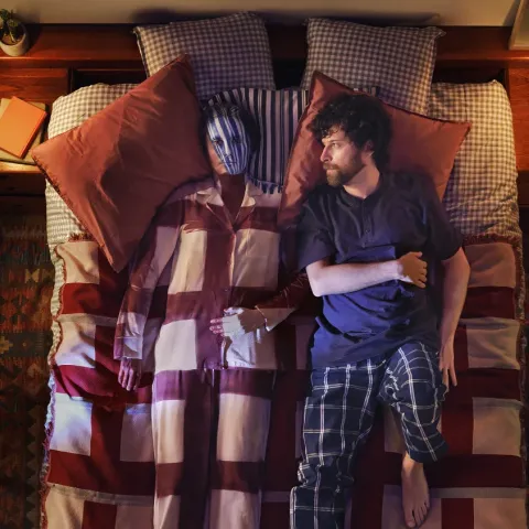 "An overhead shot of a couple lying on a bed, shot from above. A bearded man in plaid pyjama bottoms and a dark t-shirt lies beside a semi-transparent, ghostly female figure in striped pyjamas, visually representing the invisible and disorienting effects of perimenopause. The warmly lit bedroom is styled with everyday details including books, slippers and bedside lamps on a patterned rug."