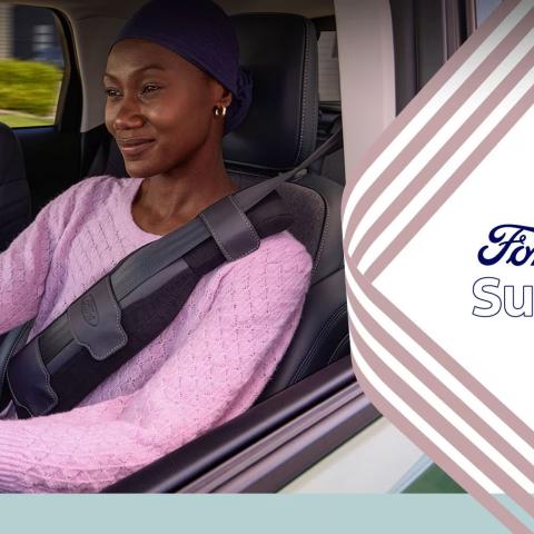 A woman wearing a pink sweater and navy head covering sits in the driver's seat of a Ford vehicle, smiling confidently while wearing the Ford SupportBelt™ — a padded seatbelt cushion positioned across her chest and shoulder.