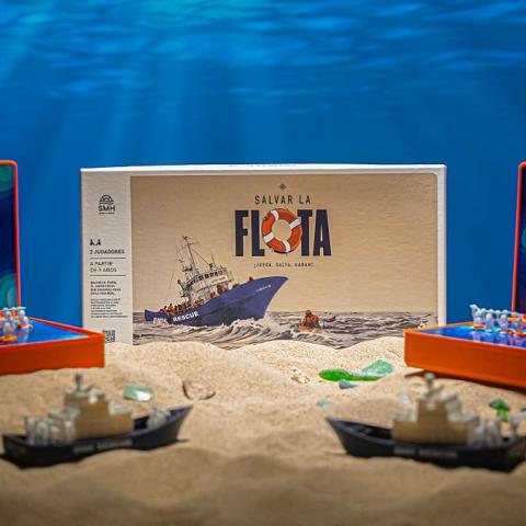 The "Salvar la Flota" board game by SMH displayed on a sandy surface with an ocean backdrop, showing the retail box flanked by two open orange game boards with playing pieces and miniature rescue ship figurines in the foreground.