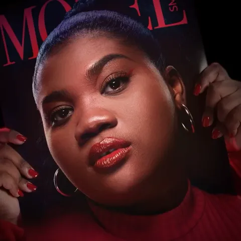 Woman holding a magazine in front of her face which looks exactly like her. The woman is a black female wearing red lipstick.
