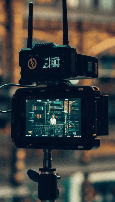 Professional video camera filming a person seated in a warmly lit industrial-style interior, viewed through the camera monitor with cinematic depth of field.