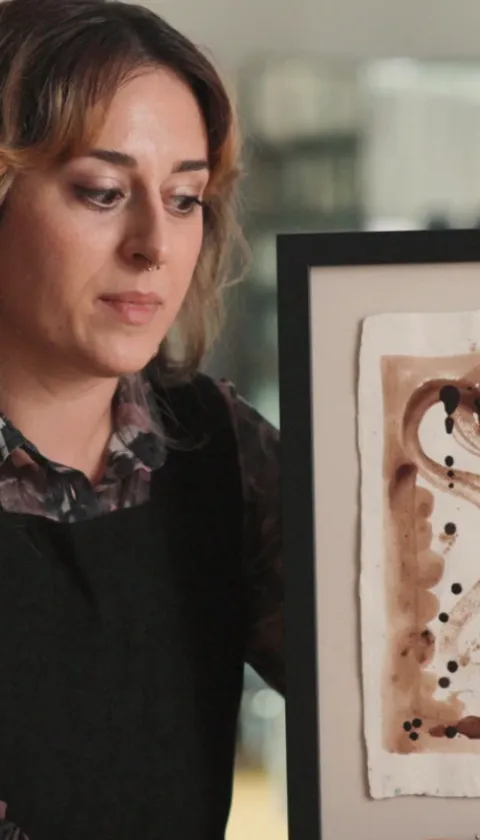 "A still from the Art's Missing Period documentary, featuring an artist holding and examining a framed abstract artwork created using menstrual fluid, with warm brown tones and gestural brushstrokes on textured paper, in a softly lit studio setting."