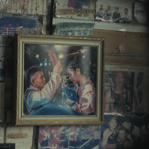 Framed photograph of a boxing trainer blessing a young female boxer before a match, displayed among family photos on a wall.