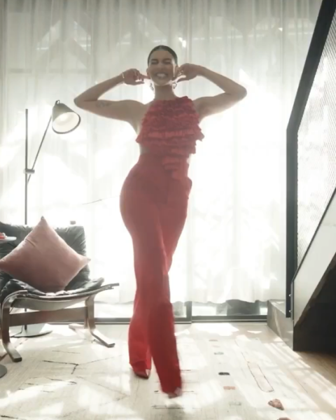 Woman in red dress standing towards camera with her hands behind her head.