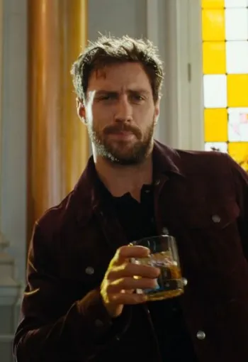 Man with brown hair drinking a glass of whiskey, stood in front of yellow windows.