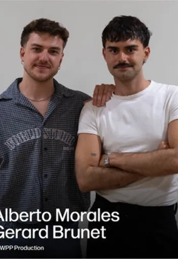 2 men standing next each other. One in a white t shirt and has a moustache. The other in a dark blue shirt with writing across the front. 