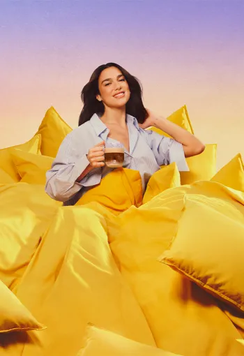 Dua Lipa sitting amongst a bed of bright yellow pillows against a soft purple backdrop