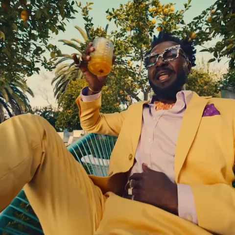 Person wearing a yellow suit sits outdoors in a garden holding a glass of orange drink, surrounded by citrus trees.